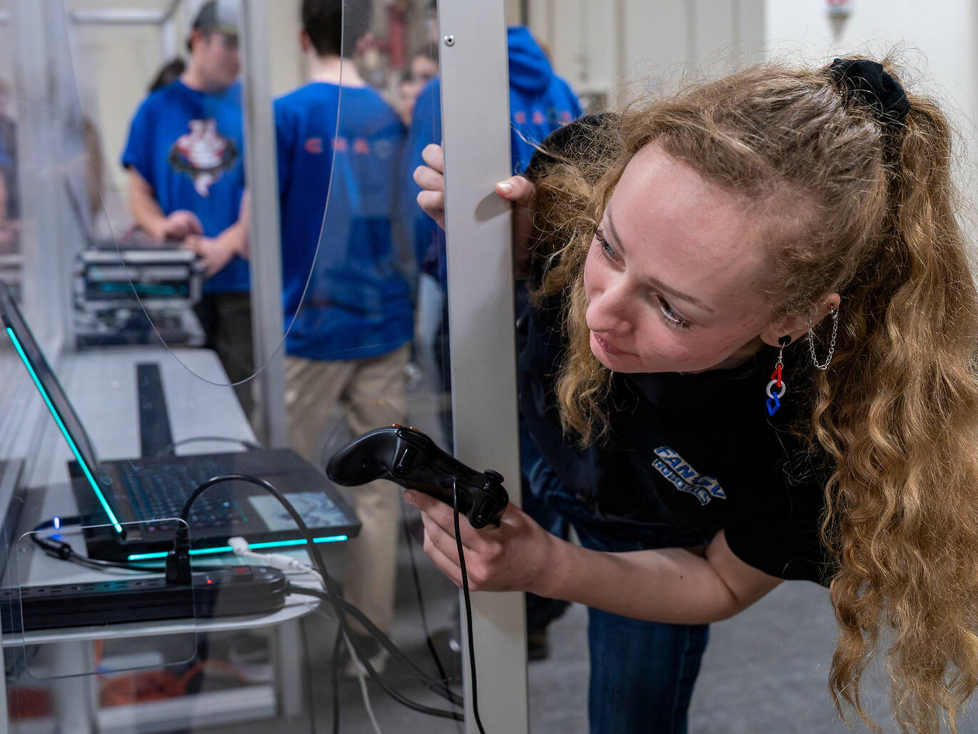 A Student with long curly hair leans over a laptop, holding a gaming controller, while others in blue shirts stand in the background.